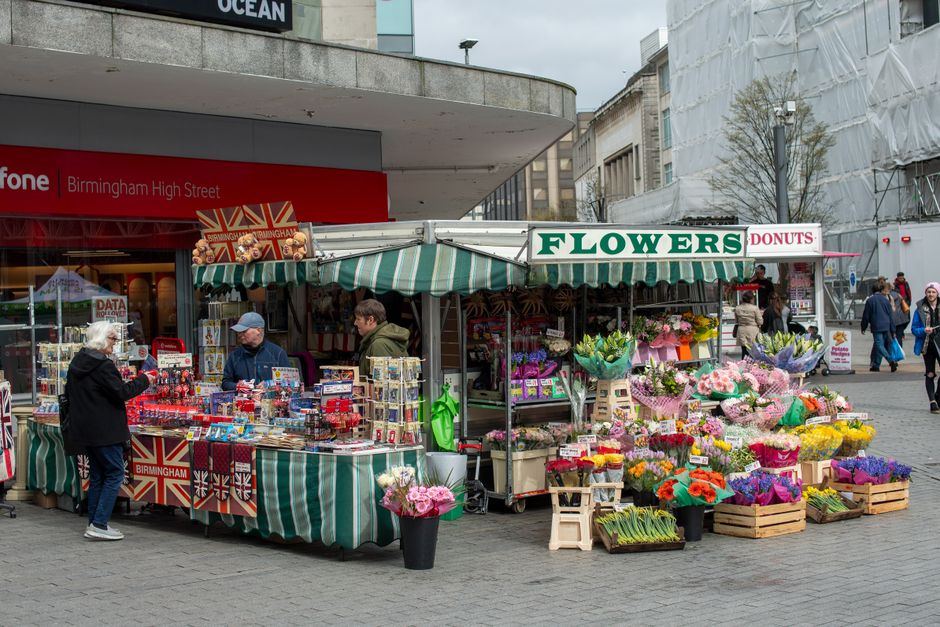 A vibrant outdoor flower stall on Birmingham High Street featuring a variety of fresh flowers arranged in wooden crates and bouquets wrapped in colorful paper. The display includes roses, daisies, tul